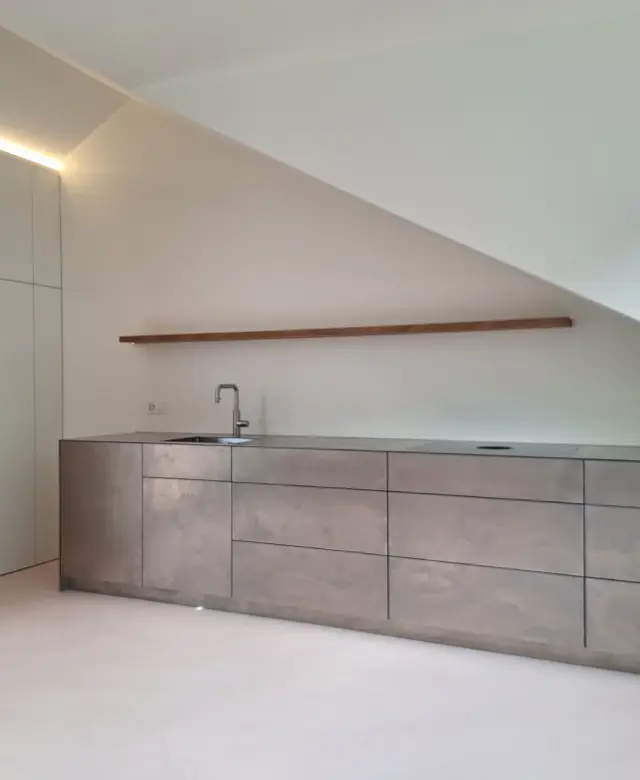 Minimalistische Wohnküche im Kanton Glarus mit griffloser Küchenzeile aus Edelstahl, rosa Spachtelboden und weißem Einbauschrank unter der Dachschräge mit LED-Lichtband von STUDIOLIVIUS.