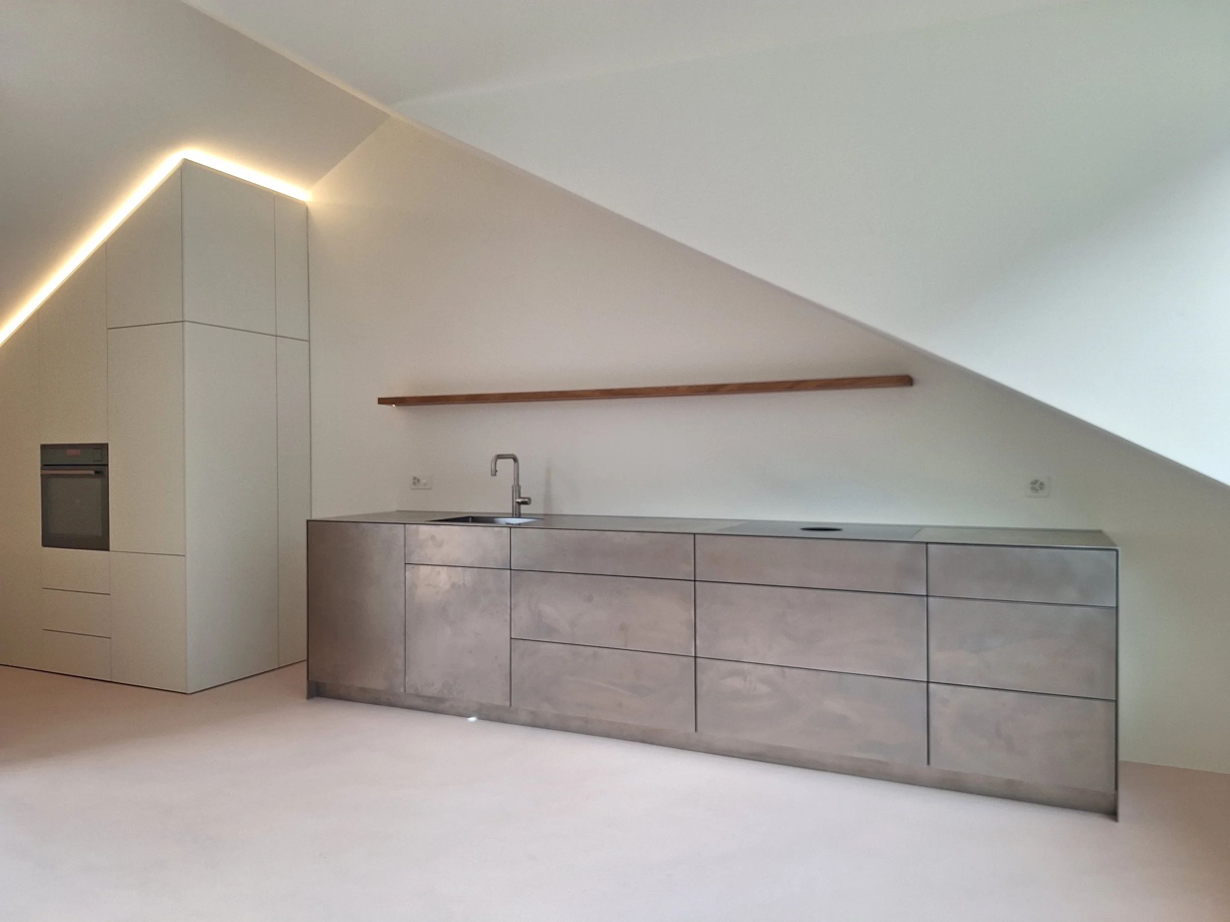 Minimalistische Wohnküche im Kanton Glarus mit griffloser Küchenzeile aus Edelstahl, rosa Spachtelboden und weißem Einbauschrank unter der Dachschräge mit LED-Lichtband von STUDIOLIVIUS.