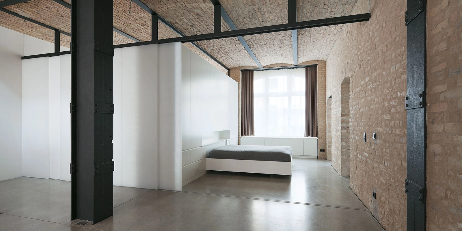 Minimalistischer Schlafbereich in einer Berliner Remise mit maßgefertigtem weißen Bett, integrierter Schrankwand, historischen Kappendecken und massiven schwarzen Stahlstützen von STUDIOLIVIUS.