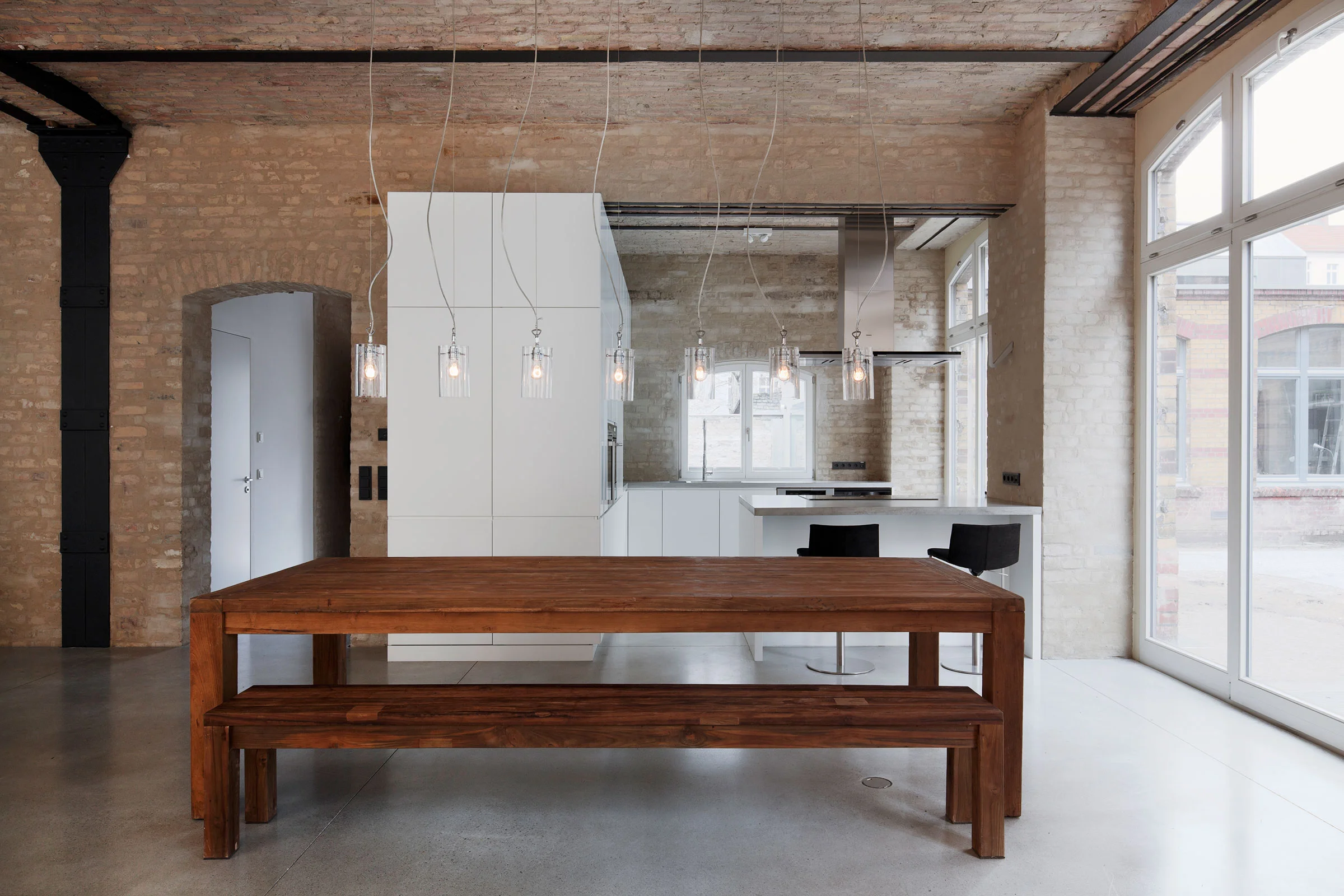 Großzügiger Essbereich im Loft mit massivem Holz-Esstisch, hängenden Industrieleuchten und einer maßgeschneiderten, grifflosen Einbauküche vor historischem Backsteinmauerwerk von STUDIOLIVIUS.