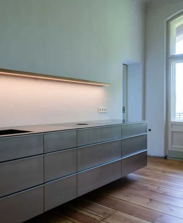Raumansicht einer schwebenden Küche aus Edelstahl mit einem beleuchteten Regalbrett in einer Berliner Altbauwonung von STUDIOLIVIUS